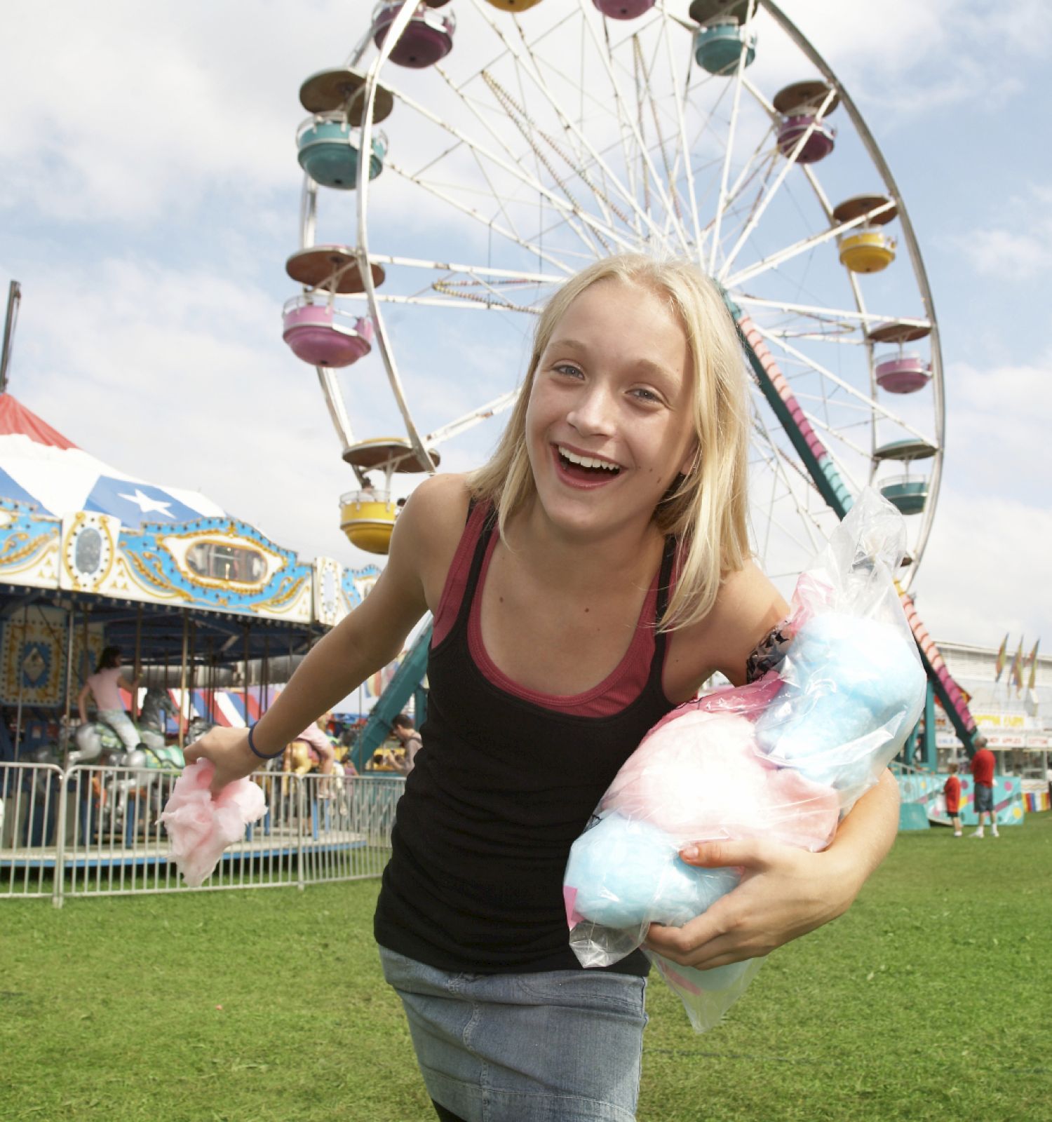 A girl smiles while holding cotton candy at an amusement park, with a Ferris wheel and carousel in the background.