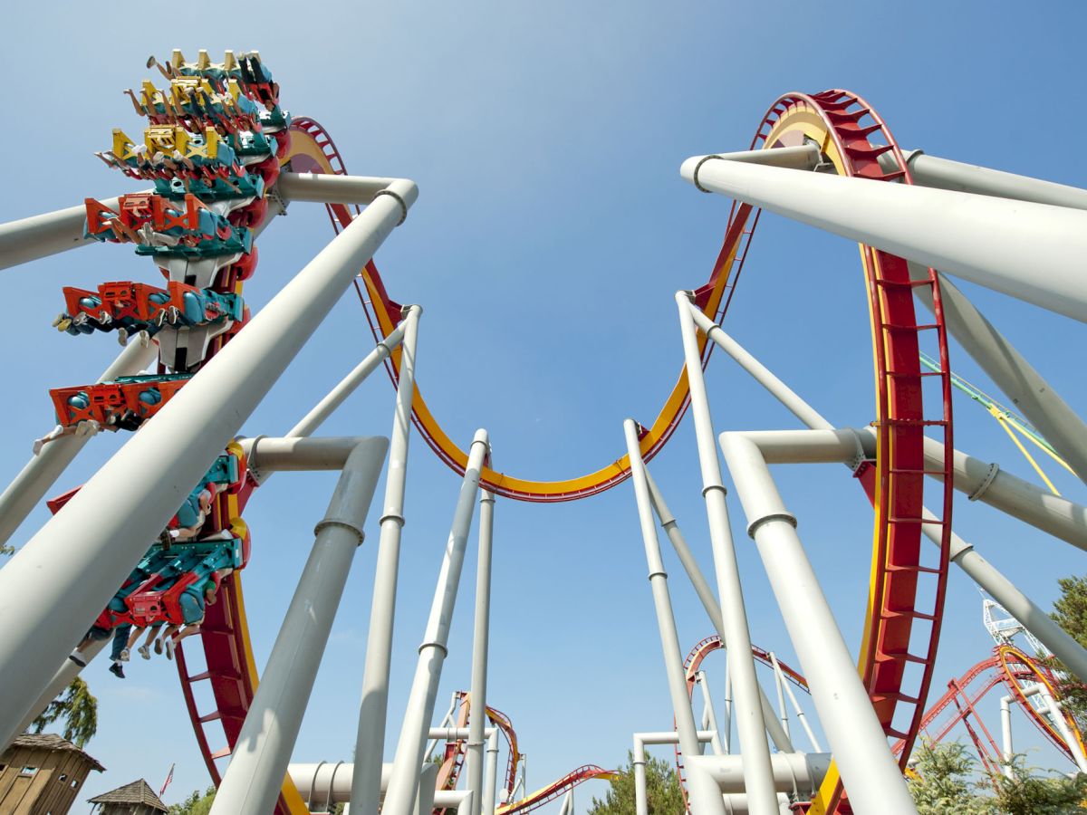 The image shows a roller coaster with red and yellow tracks, numerous loops and turns, and riders hanging down, under a clear blue sky.