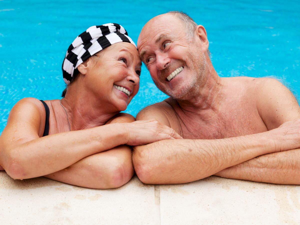 A smiling elderly couple in swimwear is leaning on the edge of a swimming pool, gazing at each other happily, enjoying their time.