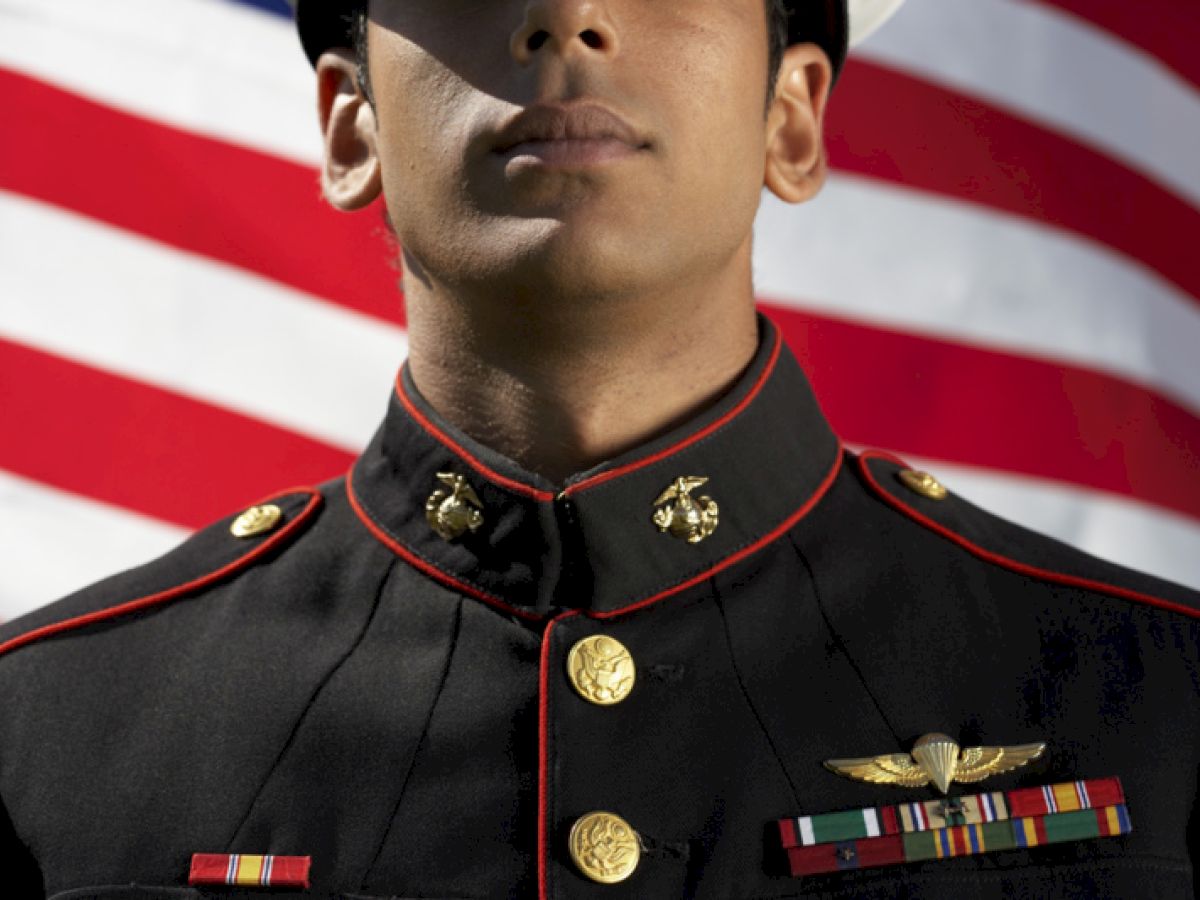 A person wearing a military uniform stands in front of an American flag, displaying various service medals and insignia on their attire.