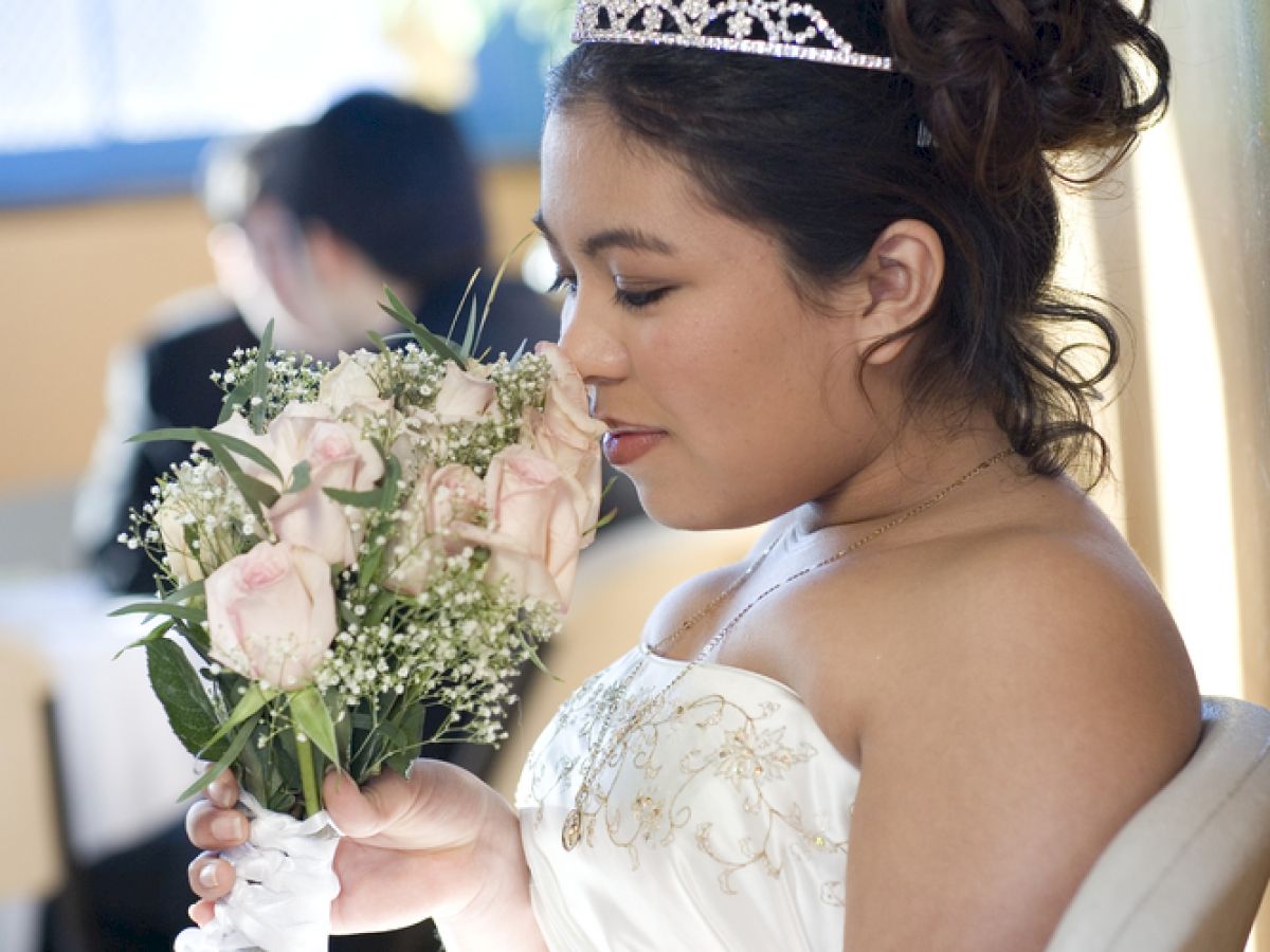 The image shows a bride wearing a tiara and holding a bouquet of flowers, sitting on a chair and looking down at her bouquet.