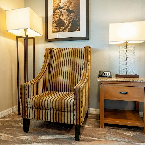 A cozy room with a striped armchair, two lamps, a bedside table, and a framed picture on the wall above the chair, ending the sentence.