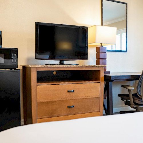 A hotel room features a TV, microwave, and mini-fridge on a wooden dresser, a lamp, a mirror, and an office chair by a desk.