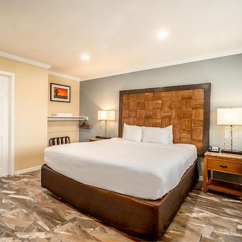 A neatly arranged bedroom with a large bed, two bedside lamps, wooden flooring, a door, a framed picture, and a wall-mounted shelf.