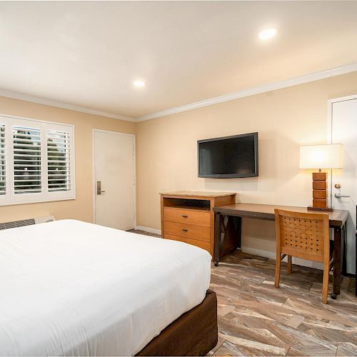 A neatly arranged hotel room featuring a bed, a flat-screen TV on the wall, a desk with a chair, a lamp, a mini fridge, and a coffee maker.