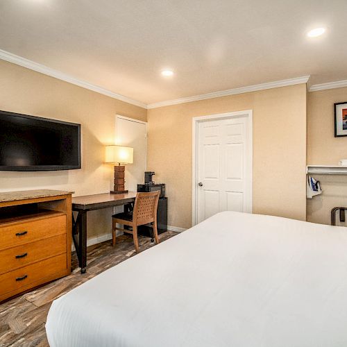 The image shows a cozy hotel room with a bed, TV, desk, lamp, and a painting on the wall next to an open closet area.