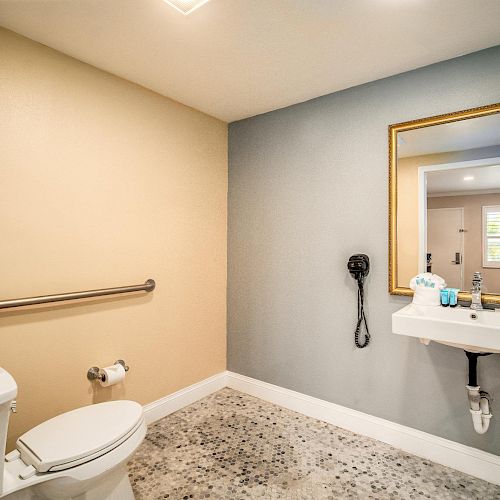 A bathroom with a toilet, a wall-mounted sink, a large mirror, a towel rack, and a hairdryer on the wall, with tiled floors and beige and gray walls.