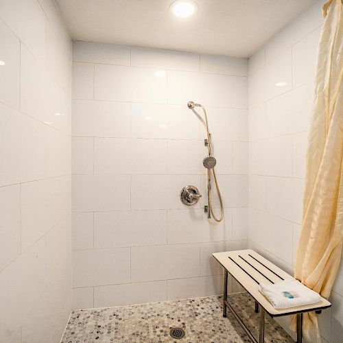 This image shows a tiled walk-in shower with a bench, a handheld showerhead, a white curtain, and a towel on the bench.