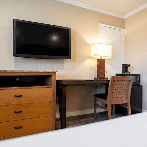 A hotel room features a wall-mounted TV, a wooden dresser, a writing desk, a chair, a lamp, and a coffee maker on a mini-fridge, ending the sentence.