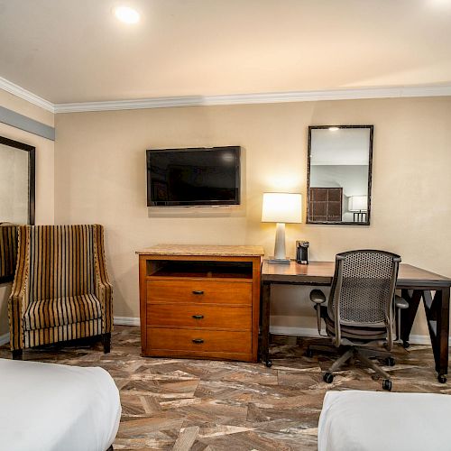 A hotel room features a wall-mounted TV, striped chair, wooden dresser, desk with lamp, mirror, and an office chair on a wood-patterned floor.