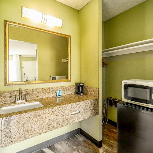 A room with a sink, mirror, hairdryer, coffee maker, microwave, mini fridge, and an open closet on green walls and wooden flooring.