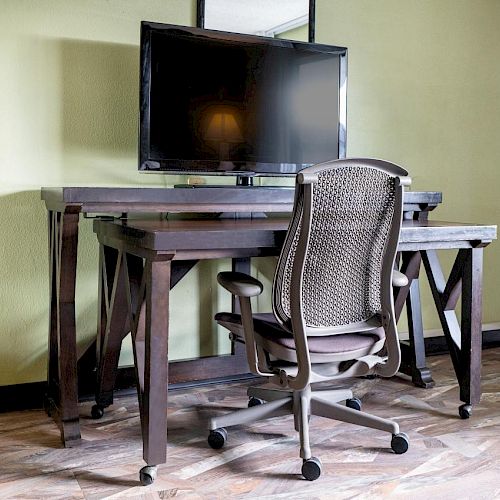 An office setup with a dark wooden desk, a mesh swivel chair, and a large monitor mounted on the desk. The room has hardwood flooring.