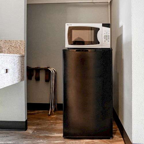 The image shows a small kitchen area with a microwave placed on top of a mini-fridge. To the left, there is a sink and a small coffee maker.