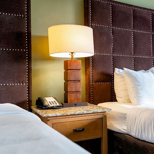 The image shows two beds with white linens and pillows, separated by a nightstand with a lamp and a telephone.