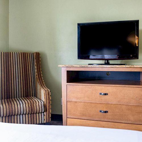 A room with a striped armchair, a wooden dresser with two drawers, and a flat-screen TV on top of the dresser.