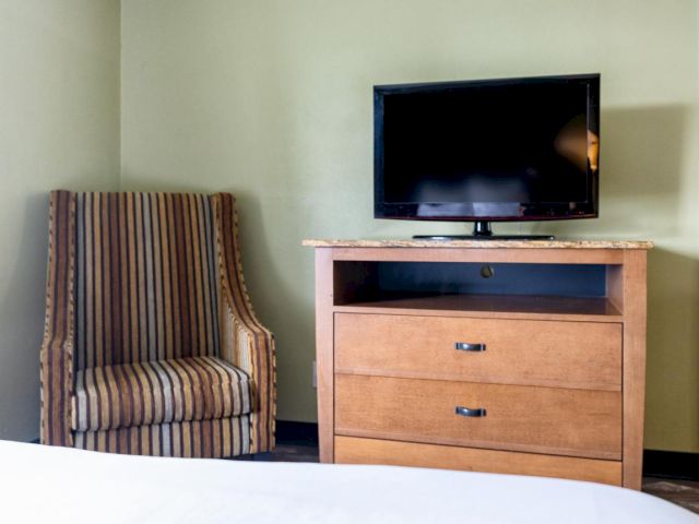 A hotel room with a striped chair, a wooden dresser with a TV on top, and a bed.