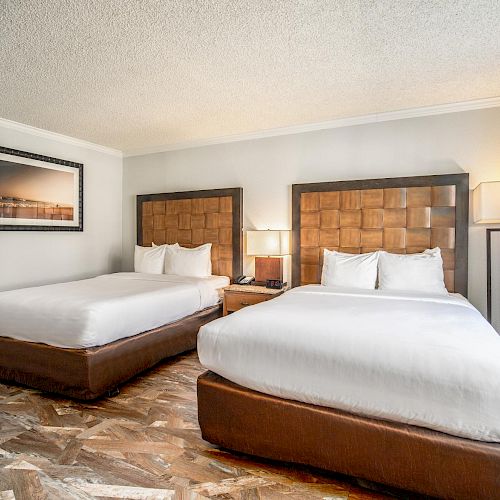 This image shows a hotel room with two double beds, a nightstand with a lamp between them, a framed picture on the wall, and a window with curtains.