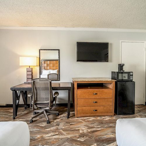 A hotel room features a desk, a chair, a lamp, a wall-mounted TV, a mini-fridge, a microwave, and an ironing board against the wall.