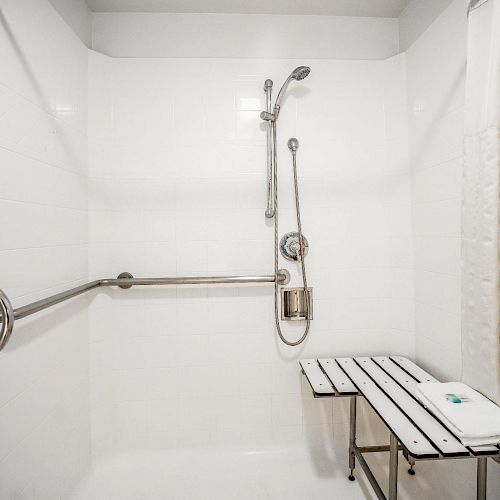 This image shows an accessible shower with a fold-down bench, grab bars, and a handheld showerhead. Towels are hung on a rail to the left of the bench.