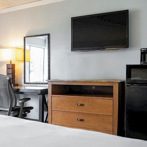 A hotel room with a desk, chair, lamp, mirror, TV mounted on the wall, and a cabinet with a microwave and mini-fridge.