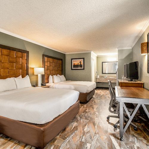 This image shows a hotel room with two beds, a TV on a desk, a chair, bedside lamps, and a wall mirror, decorated in a modern style.