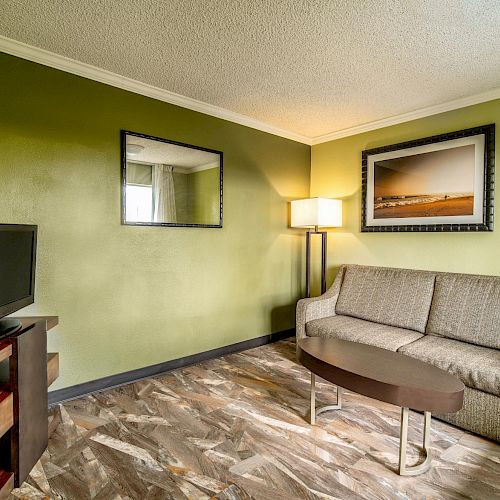 A living room with green walls, a beige sofa, a TV on a wooden stand, a mirror, a framed picture, a floor lamp, and a coffee table.