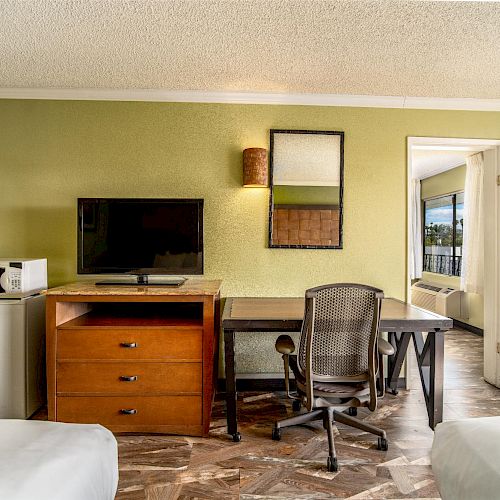 A hotel room with a TV, microwave, mini-fridge, a desk, and an office chair against a green wall, with a doorway to another room.