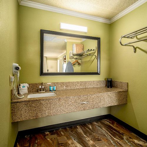 This image shows a hotel bathroom area with a large mirror above a granite countertop, a hairdryer, toiletries, and a coat rack on a green wall.