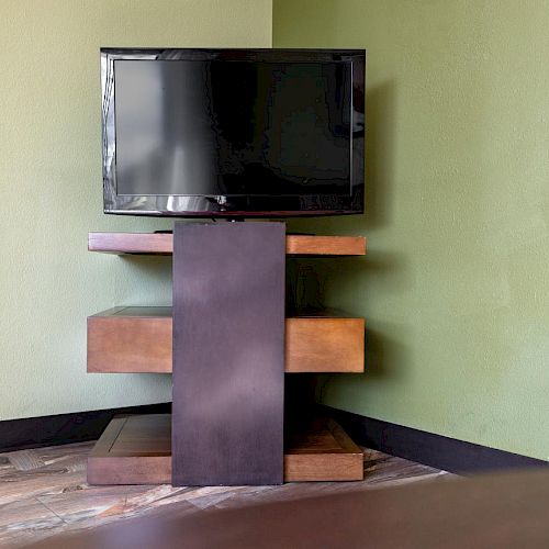 A flat-screen TV on a modern wooden stand, placed in the corner of a room with green walls and tile flooring.