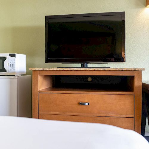 The image shows a TV on a wooden stand, a microwave on a mini-fridge, a table, and part of a bed in a room.