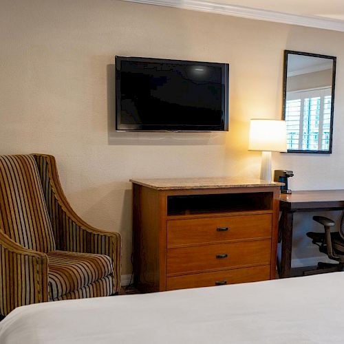 A cozy hotel room features a striped armchair, a wall-mounted TV, a wooden dresser, and a work desk with a chair beside a lamp and a mirror on the wall.