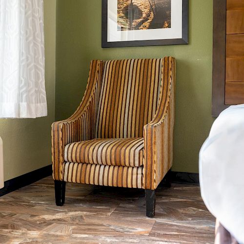 The image shows a striped armchair next to a bed in a room with green walls, a white curtain, a wall-mounted air conditioner, and framed artwork.