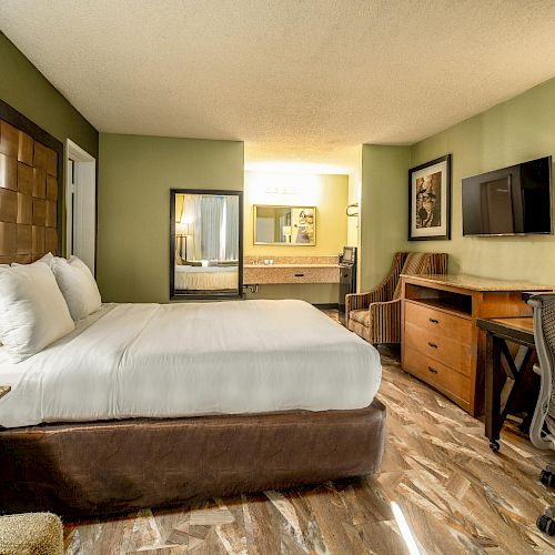 This image shows a well-lit hotel room with a large bed, a desk and chair, a flat-screen TV, and a vanity area with a mirror and sink.