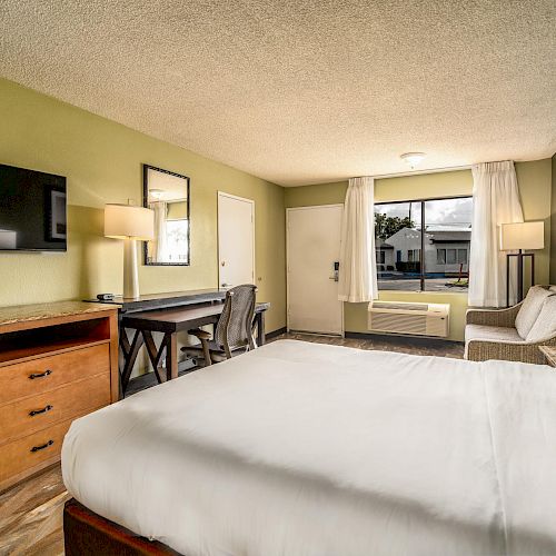 A hotel room with a bed, TV on a wooden dresser, desk and chair, sofa, and a window with curtains. The room has green and beige walls, and wood flooring.