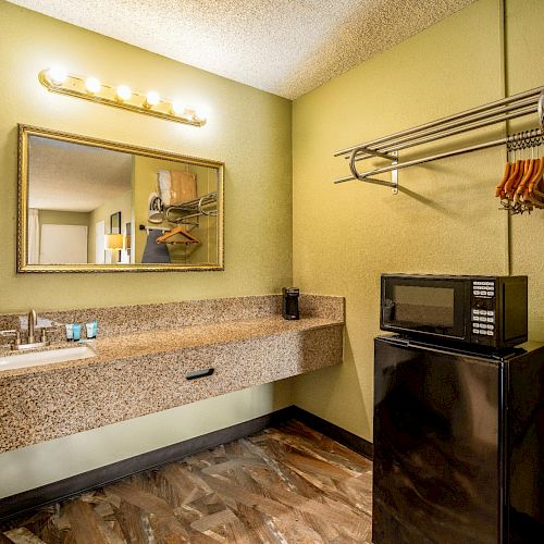 A small room with a sink, mirror, hairdryer, countertop, microwave, mini-fridge, and a clothes rack with hangers.