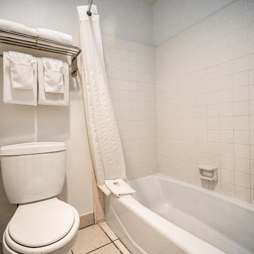 A clean bathroom with a white toilet, a bathtub with a shower curtain, and neatly folded towels on a shelf above the toilet.