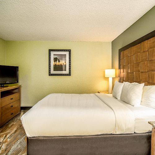 The image shows a neatly made bed in a hotel room, with a wooden headboard, TV on a wooden cabinet, artwork on the wall, and bedside lamps.