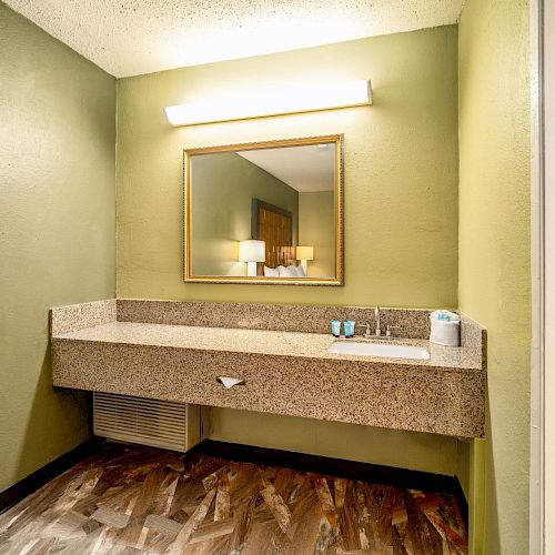 A bathroom with green walls, a large mirror, a countertop with a sink, toiletries, and a light fixture above. The floor is wood-patterned.