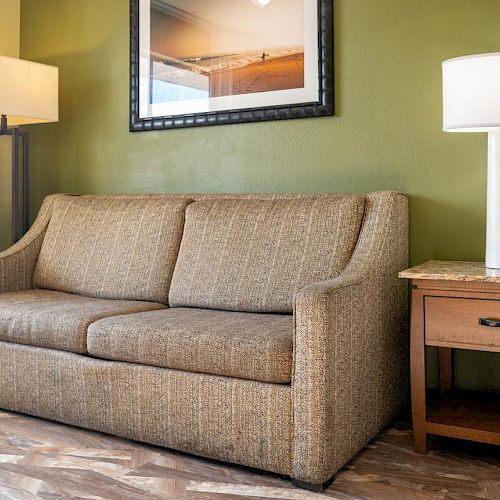 A cozy living room setup with a brown sofa, two floor lamps, a side table, and a framed photo on a green wall at the back.