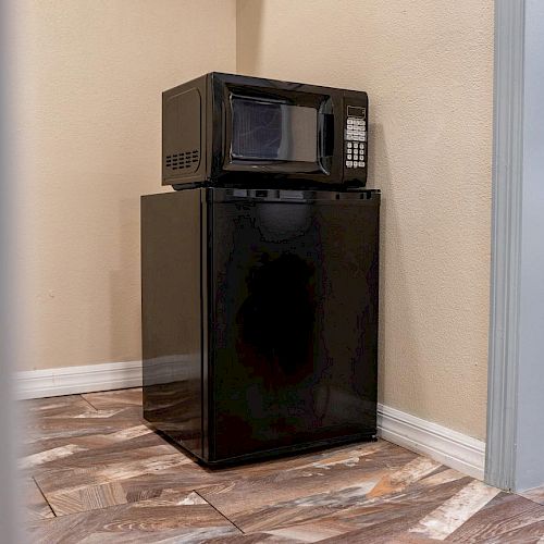 A black microwave sits atop a black mini-fridge in a small kitchenette area with wood-patterned tile flooring and neutral wall colors.