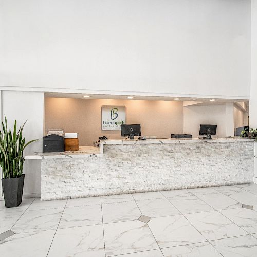 A modern, welcoming hotel reception area with a marble counter, two potted plants, computers, and a logo on the wall.