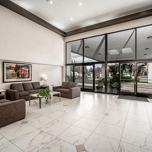 A modern, spacious lobby with gray sofas, coffee tables, wall art, and large glass doors leading outside, surrounded by white marble flooring and potted plants.