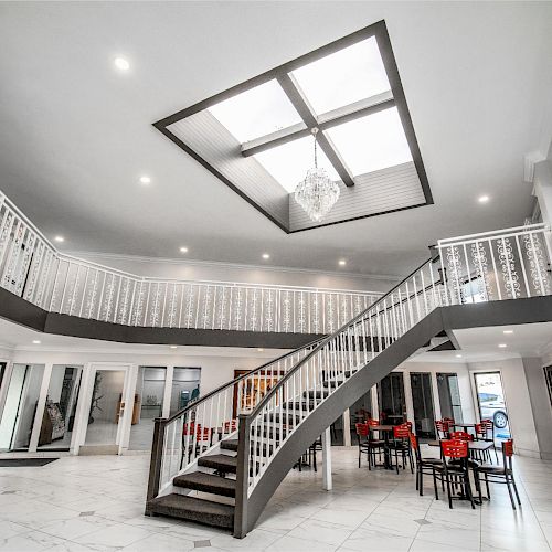 The image shows an indoor space with a grand staircase, white railings, a skylight, and tables with red chairs, likely a lobby or reception area.