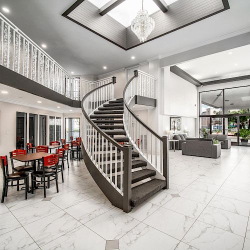 A spacious, modern lobby with a grand spiral staircase, marble floors, a seating area with red chairs and tables, and large windows letting in natural light.