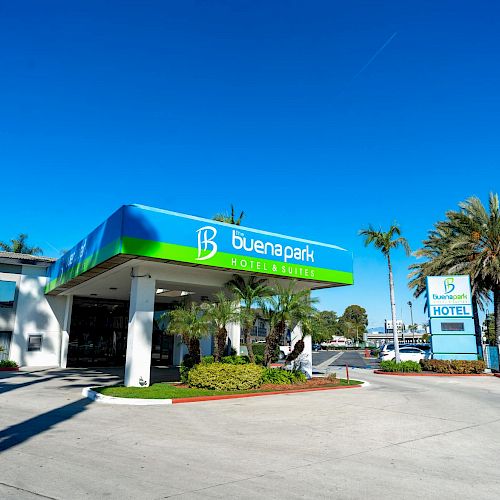 The image shows the entrance of a hotel named "Buena Park," with a canopy, landscaping, and palm trees around. The sky is clear and blue.