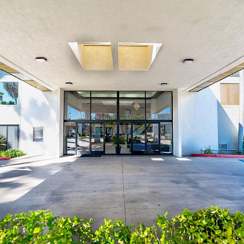 The image shows the entrance of a modern building with large glass doors, overhang, and greenery in the foreground. The weather appears sunny.