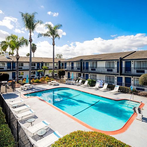 A residential complex with a central swimming pool, surrounded by sun loungers and palm trees, with two-story buildings in the background.