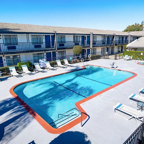 An outdoor swimming pool surrounded by lounge chairs with a two-story motel in the background on a sunny day, providing a relaxing setting.