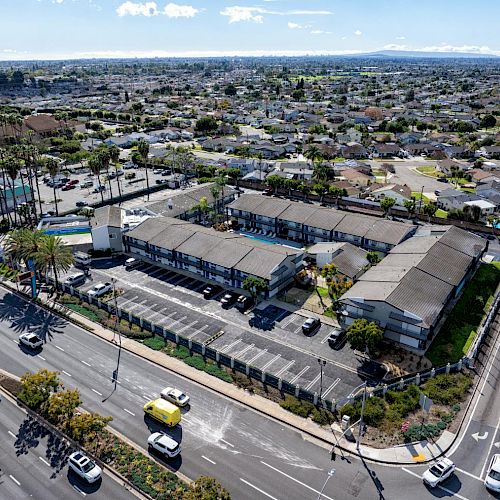 An aerial view of a suburban area featuring a large building complex with parking, surrounded by busy roads with vehicles and residential houses.