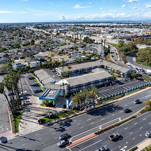 An aerial view of a cityscape featuring a busy road, buildings, and a large parking area surrounded by greenery on a sunny day.